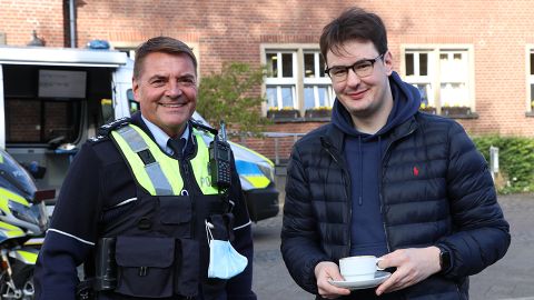 Zwei Personen, einer in Polizeiuniform stehen nebeneinander. Die rechte Person hält eine Kaffeetasse. 